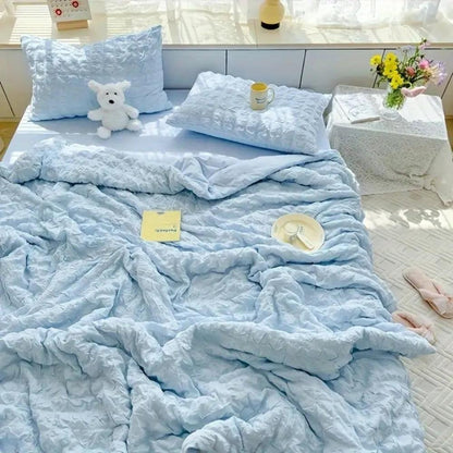 Blue textured comforter on a bed with pillows and a teddy bear in a bright room.