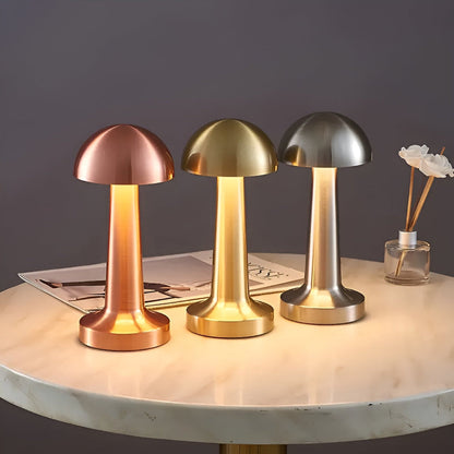 Three mushroom-shaped table lamps in bronze, gold, and silver on a marble surface with a dark background.